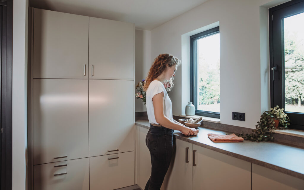 Vrouw in op maat gemaakte bijkeuken met moderne opbergkasten, werkblad en decoratieve elementen bij het raam.