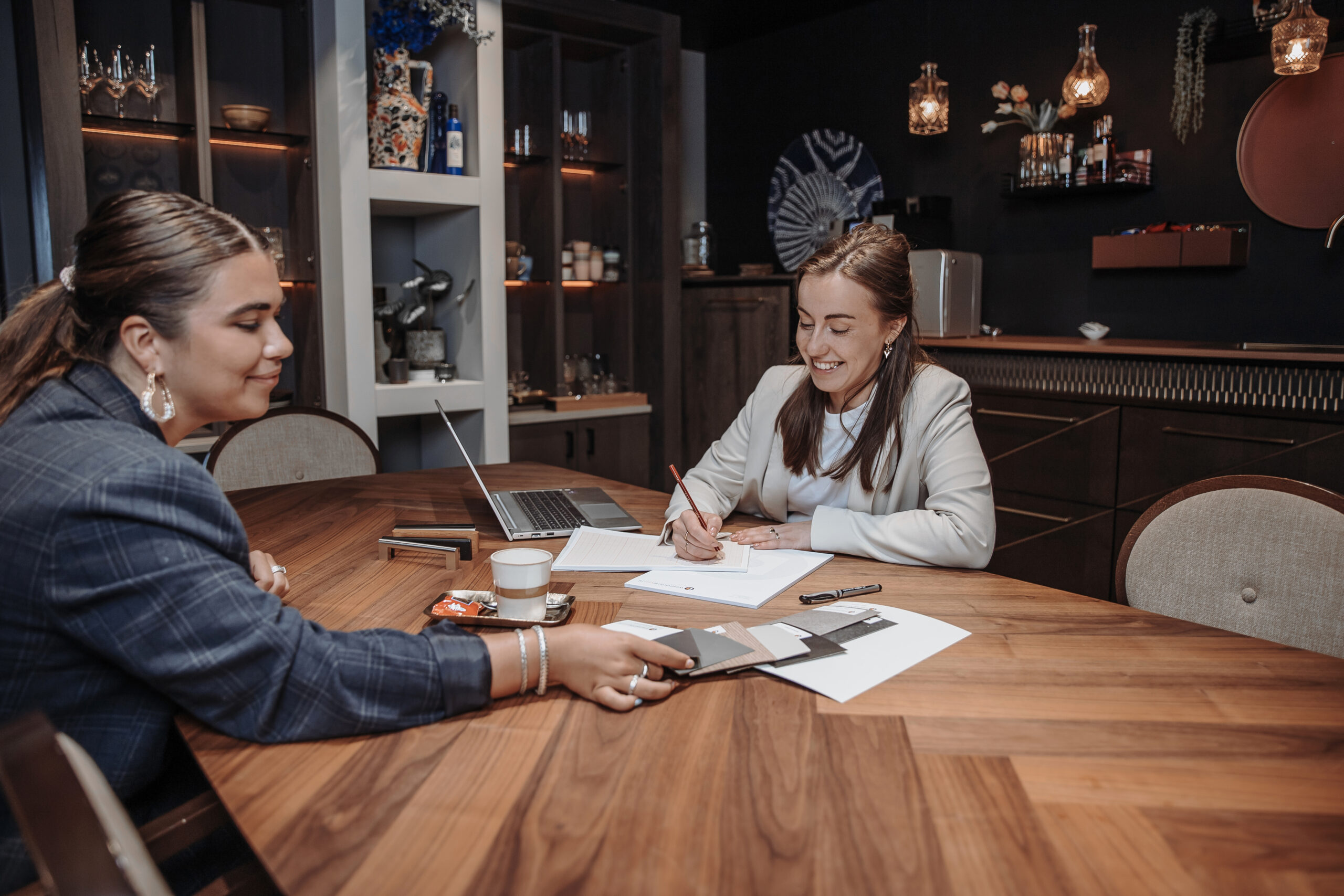 Twee vrouwen aan een tafel bekijken kleurstalen, met laptop en kopje koffie in een stijlvolle showroom.